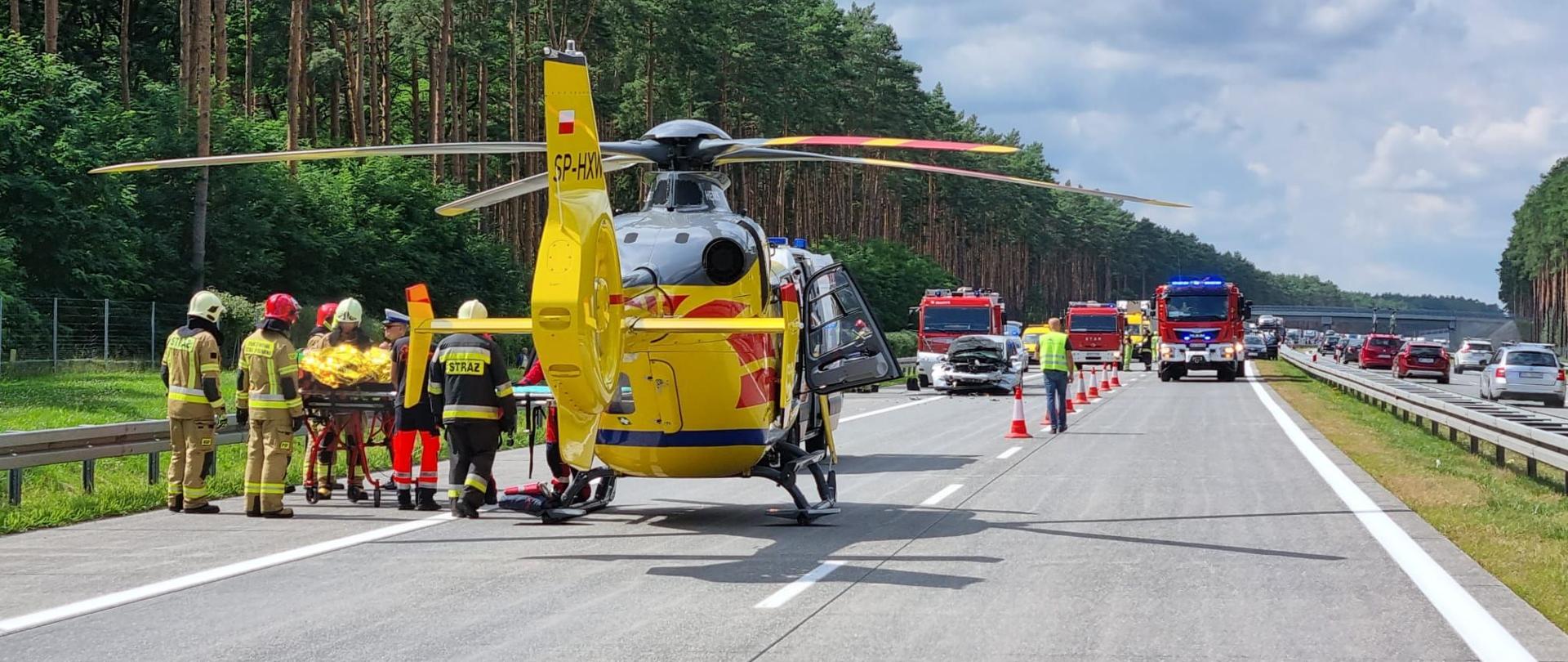 Dzień. Na autostradzie stoi śmigłowiec Lotniczego Pogotowia Ratunkowego, obok strażacy transportują na noszach osobę poszkodowaną.