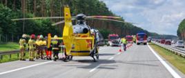 Dzień. Na autostradzie stoi śmigłowiec Lotniczego Pogotowia Ratunkowego, obok strażacy transportują na noszach osobę poszkodowaną.