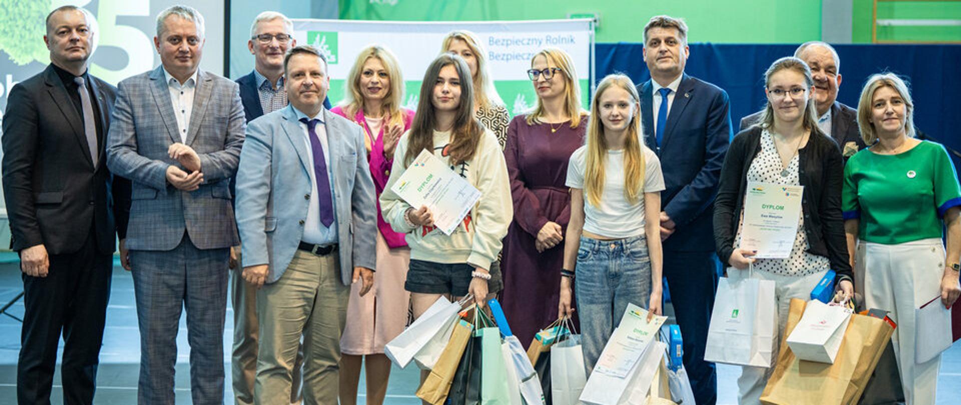 Grupa 13 osób pozuje do zdjęcia na sali gimnastycznej. W pierwszym rzędzie stoją trzy dziewczynki z licznymi torbami z nagrodami i dyplomami w rękach oraz mężczyzna i kobieta. Pozostali – czterej mężczyźni i trzy kobiety – stoją w drugim rzędzie. Za nimi jest umieszczona ścianka z logo Kasy.