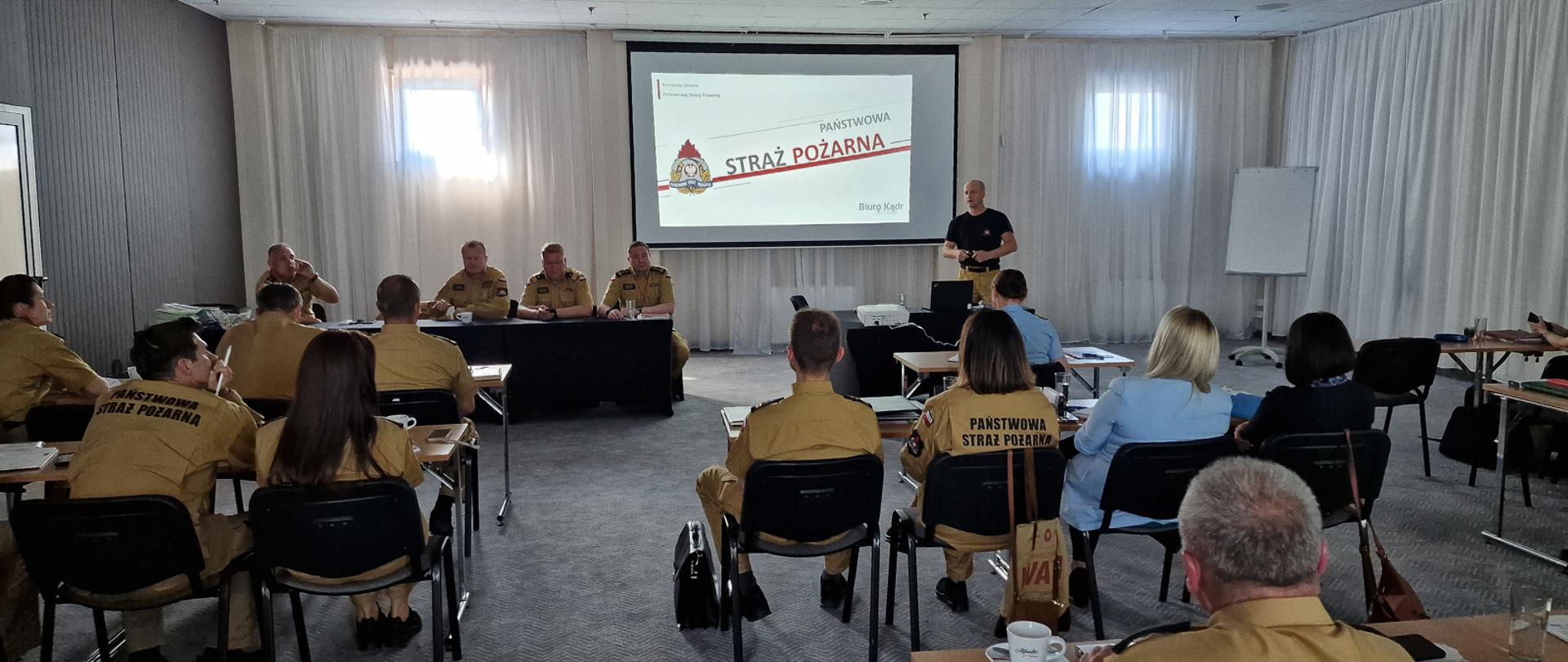 strażacy i inne osoby siedzą w sali konferencyjnej, na ekranie rzutnika wyświetlony jest slajd o treści Państwowa Straż pożarna biuro kadr