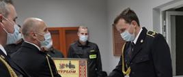 Komendant powiatowy mł. bryg. Arkadiusz Makowski ubrany w mundur galowy ze sznurem w maseczce ochronnej na twarzy wręcza pamiątkowy dyplomu z podziękowaniami za wzorową służbę od komendanta głównego nadbryg. Andrzeja Bartkowiaka strażakowi tutejszej komendy, na wprost w głębi trzech strażaków w pozycji zasadniczej stoją w szeregu, ubrani w ubranie koszarowe i maseczkach ochronnych na twarzy
