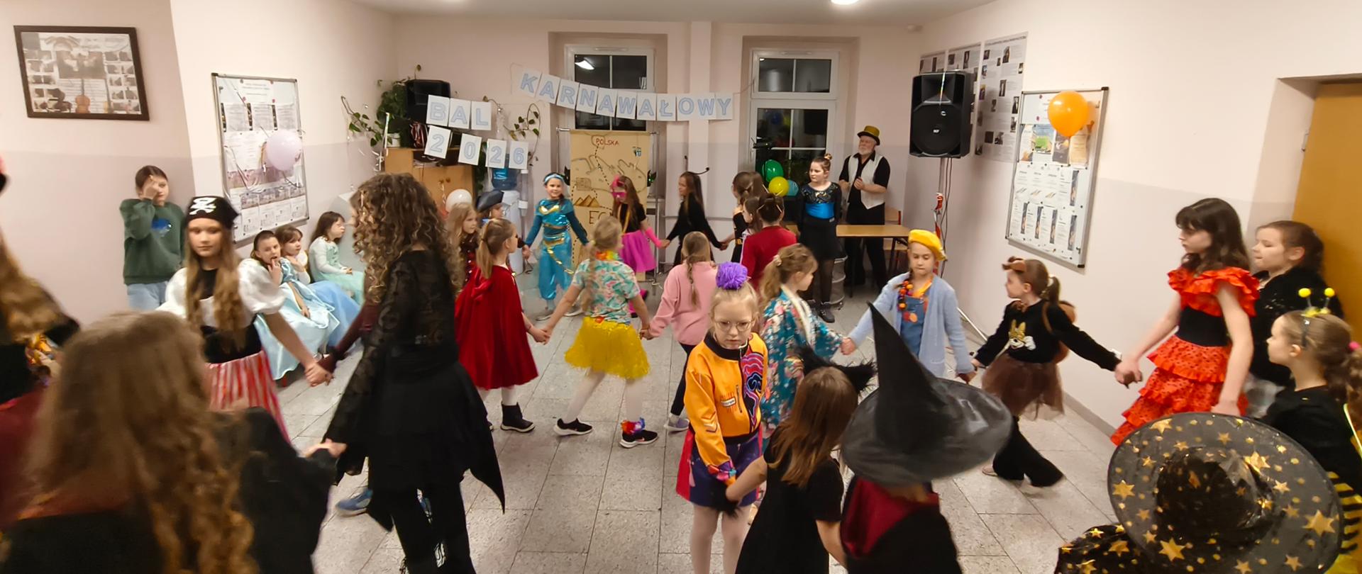 Dzieci przebrane za piratów, wróżki i inne postacie tańczą w dużym kole, trzymając się za ręce. Sala jest jasno oświetlona, a w głębi widać sprzęt nagłaśniający oraz karnawałowe napisy.