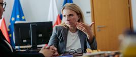Wiceministra funduszy i polityki regionalnej Monika Sikora, wiceministra siedzi przy stole, za jej plecami flagi Polski i UE