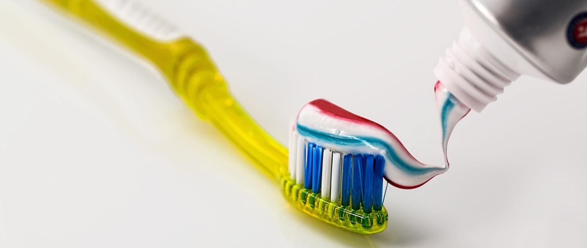 Na biały tle żółta szczoteczka z czerwono-niebiesko-zieloną pastą