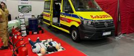 Stoisko Państwowej Straży Pożarnej na którym stoi ambulans PSP w kolorze żółtym, obok którego przygotowane są manekiny do treningu pierwszej pomocy oraz gaśnice
