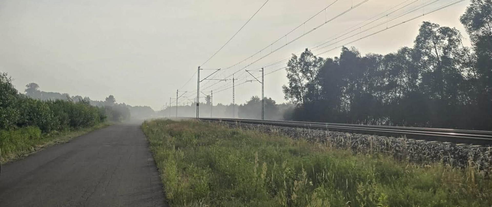 Na fotografii po lewej stronie droga asfaltowa. po prawej stronie tory kolejowe i teren zielony przy torach. W oddali widać unoszący się dym.