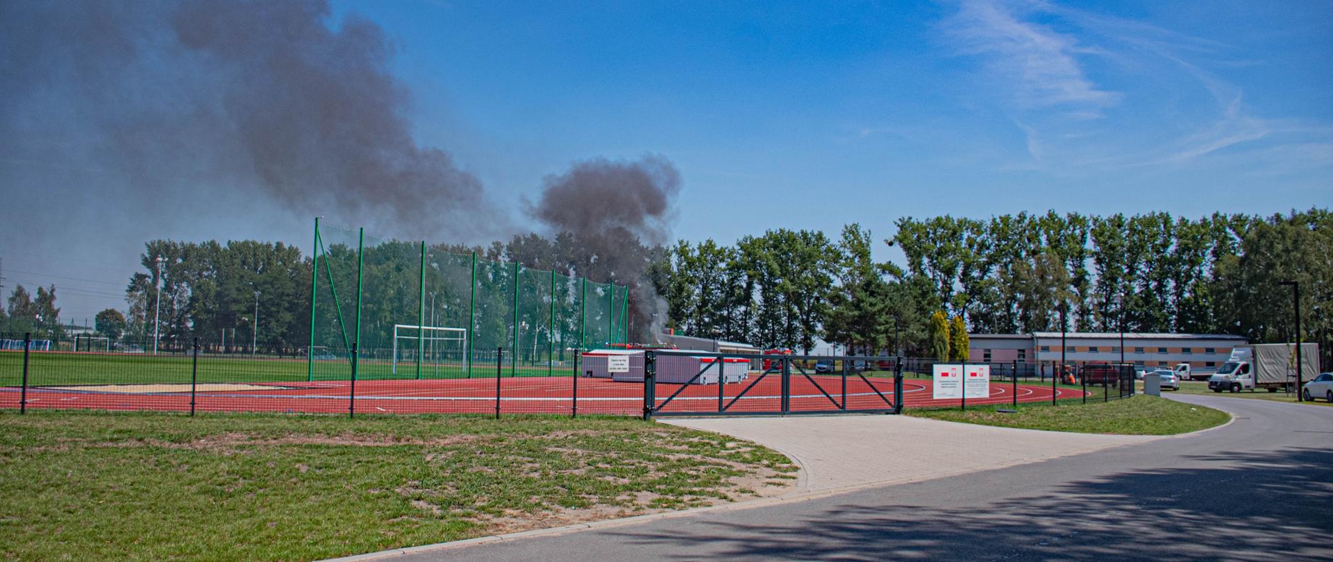 W oddali widać zadymienie. Pali się samochód zaparkowany na terenie kompleksu sportowego. Widok na boisko piłkarskie, bieżnie lekkoatletyczną oraz budynki należące do kompleksu