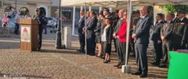 Strażacy i zaproszeni goście stoją pod namiotem zebrani na płycie rynku. jeden z strażaków przemawia przy mównicy. W tle ludzie przechodzący ulicą.