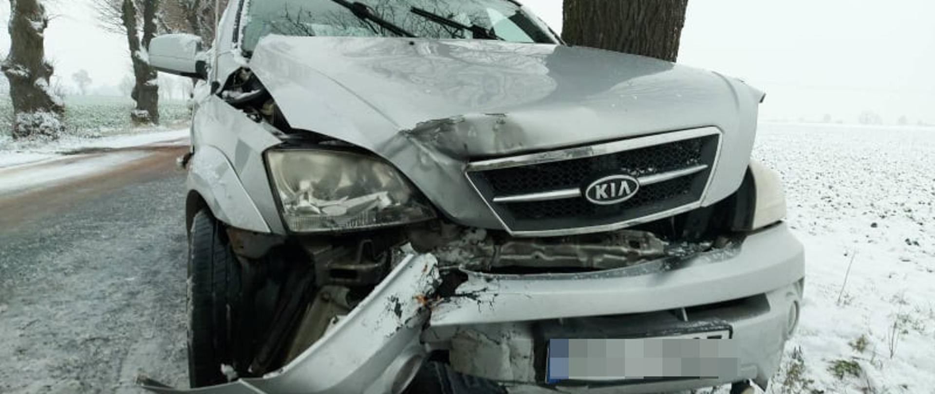 Kolizja pod Jutrosinem. Droga powiatowa Jutrosin-Dłoń. Na oblodzonej i ośnieżonej jezdni, przy drzewie stoi samochód osobowy koloru jasnego, który uległ kolizji. Auto ma rozbity przód. Po obu stronach jezdni rosną drzewa. Wokół pola pokryte śniegiem.