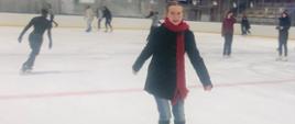 Zdjęcie uśmiechniętej dziewczyny na lodowisku. W tle widać inne osoby korzystające z jazdy na łyżwach.