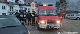 Kolorowa fotografia przedstawia widok utwardzonego placu parkingowego, na którym stoi 5 strażaków OSP w umundurowaniu, w maseczkach ochronnych i rękawiczkach ochronnych. W rękach trzymają roznoszone ulotki informacyjne. Z prawej strony obok strażaków widoczny czerwony pojazd bojowy straży pożarnej. W tle widoczne samochody osobowe na parkingu, część elewacji budynku wielorodzinnego oraz drzewa.