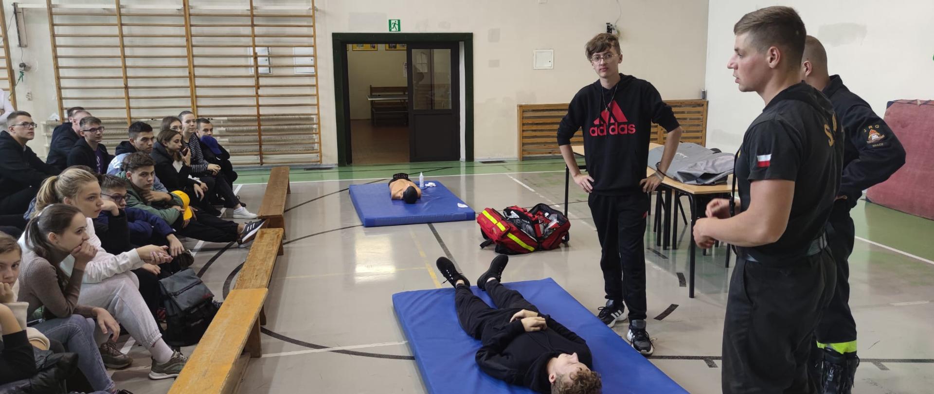 Zdjęcie przedstawia strażaka prowadzącego pogadankę. Na materacu leży młody mężczyzna w roli pozoranta. Wszystkiemu przygląda się siedząca na ławeczkach młodzież.