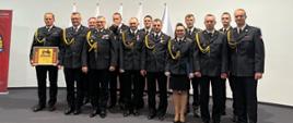 Dwa szeregi strażaków w galowych czarnych mundurach, którzy stoją w szeregu na podwyższeniu w jasnym pomieszczeniu. W tle na ścianie wisi krzyż i godło polskie. Kliknięcie powoduje powiększenie zdjęcia.