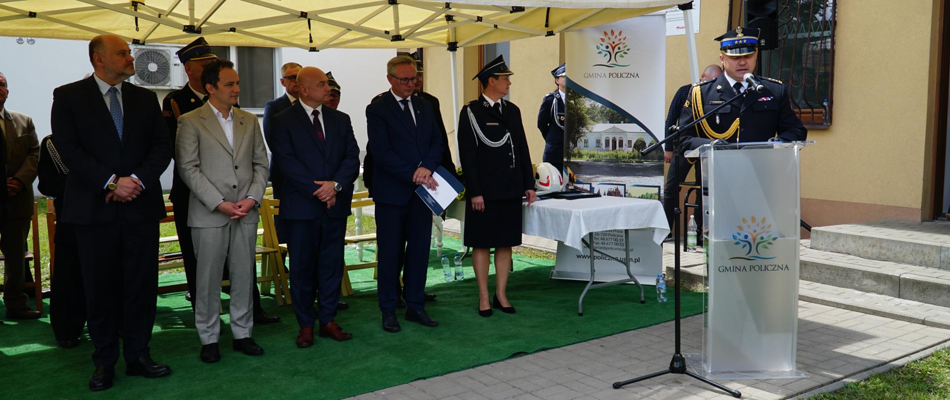 Oficjalne uroczystości przed budynkiem z napisem „OSP CZARNOLAS”. Po prawej stronie, przy przezroczystej mównicy z logo „GMINA POLICZNA”, stoi strażak w galowym mundurze, który przemawia do mikrofonu. Po lewej stronie, pod białym namiotem ogrodowym rozstawionym na sztucznej trawie, stoi grupa zaproszonych gości – mężczyźni w garniturach oraz funkcjonariusze w mundurach. W tle widoczny jest roll-up reklamowy Gminy Policzna oraz stolik nakryty białym obrusem, na którym leży strażacki kask.
