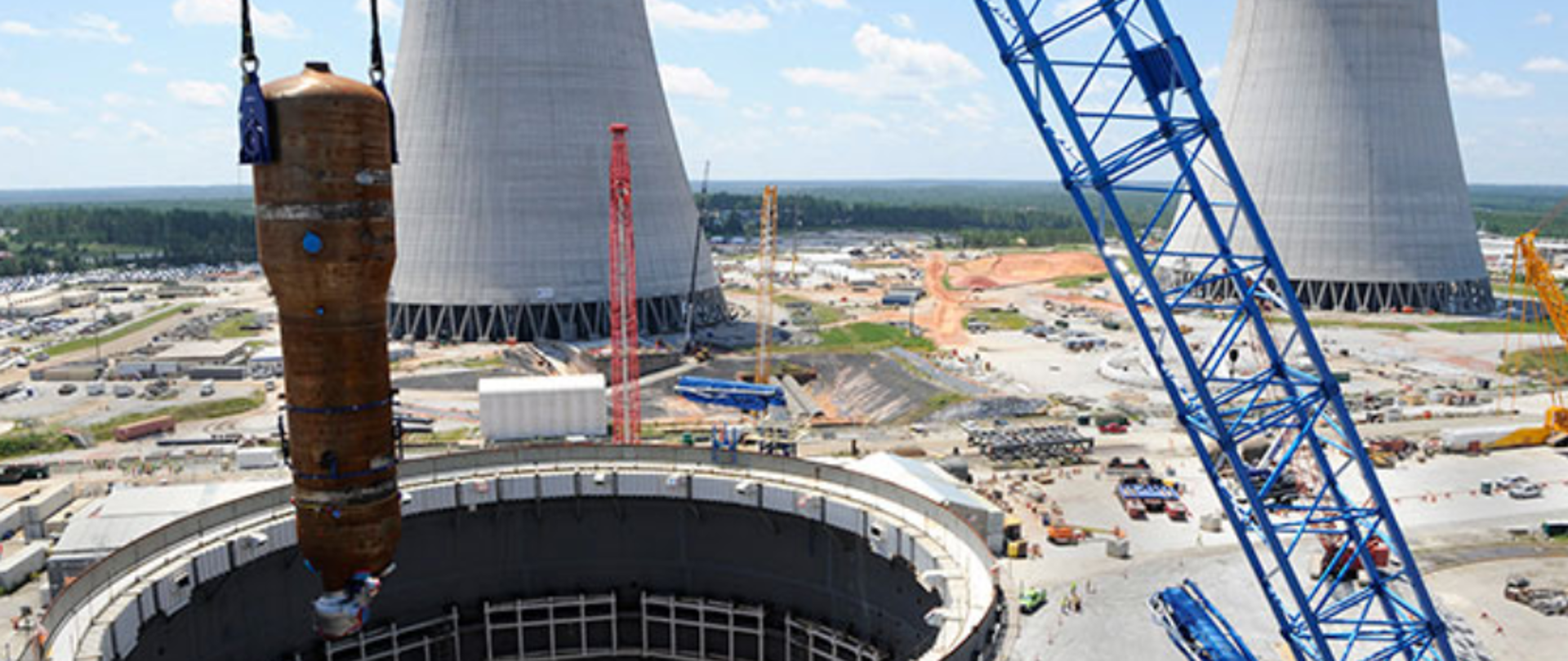 Budowa elektrowni jądrowej Vogtle 3 i 4. Niebieskie ramię dźwigu umieszcza element w budynku reaktora jądrowego. Obok plac budowy - maszyny budowlane. W tle dwa, szare kominy chłodzące i zielony las.