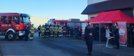 Strażacy stoją w szeregu na tle pojazdów strażackich, na pierwszym tle przemawia strażak. Przy remizie strażąckiej stoi namiot z osobami cywilnymi i strażakami.
