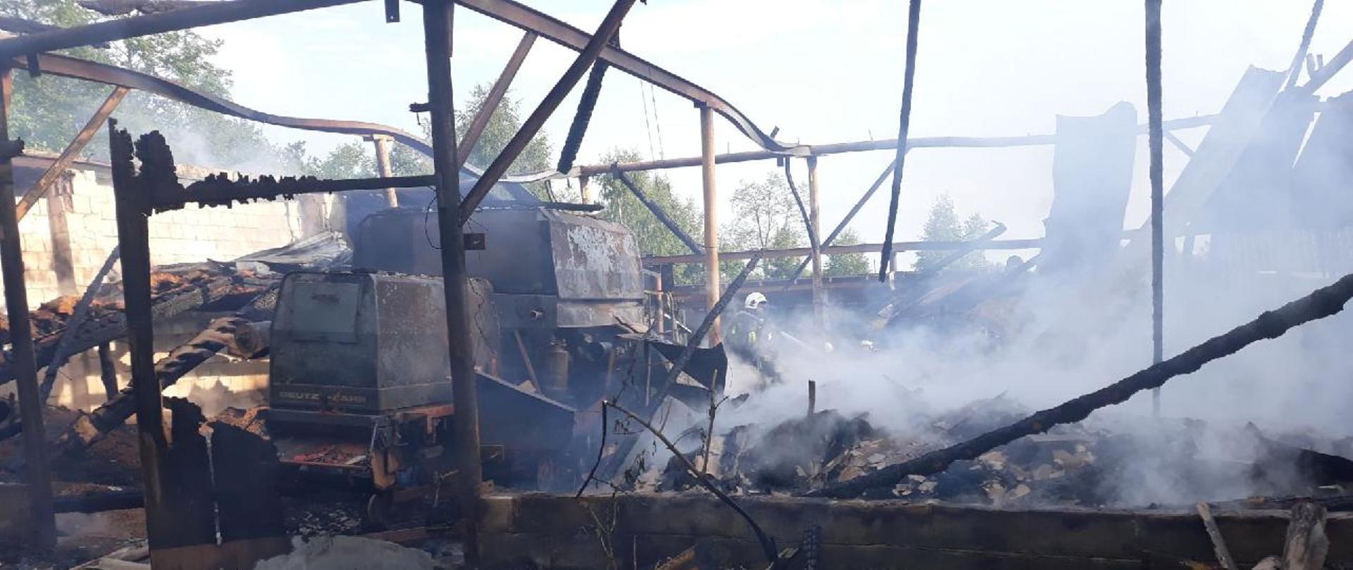 Zdjęcie przedstawia pogorzelisko po pożarze budynku gospodarczego ze spalonymi elementami konstrukcji, bez dachu, wewnątrz spalony kombajn zbożowy, oraz zawalone części dachu, ścian oraz przechowywanych wewnątrz płodów rolnych. Widoczny unoszący się dym, w tle strażak OSP dogaszający pogorzelisko prądem wody.