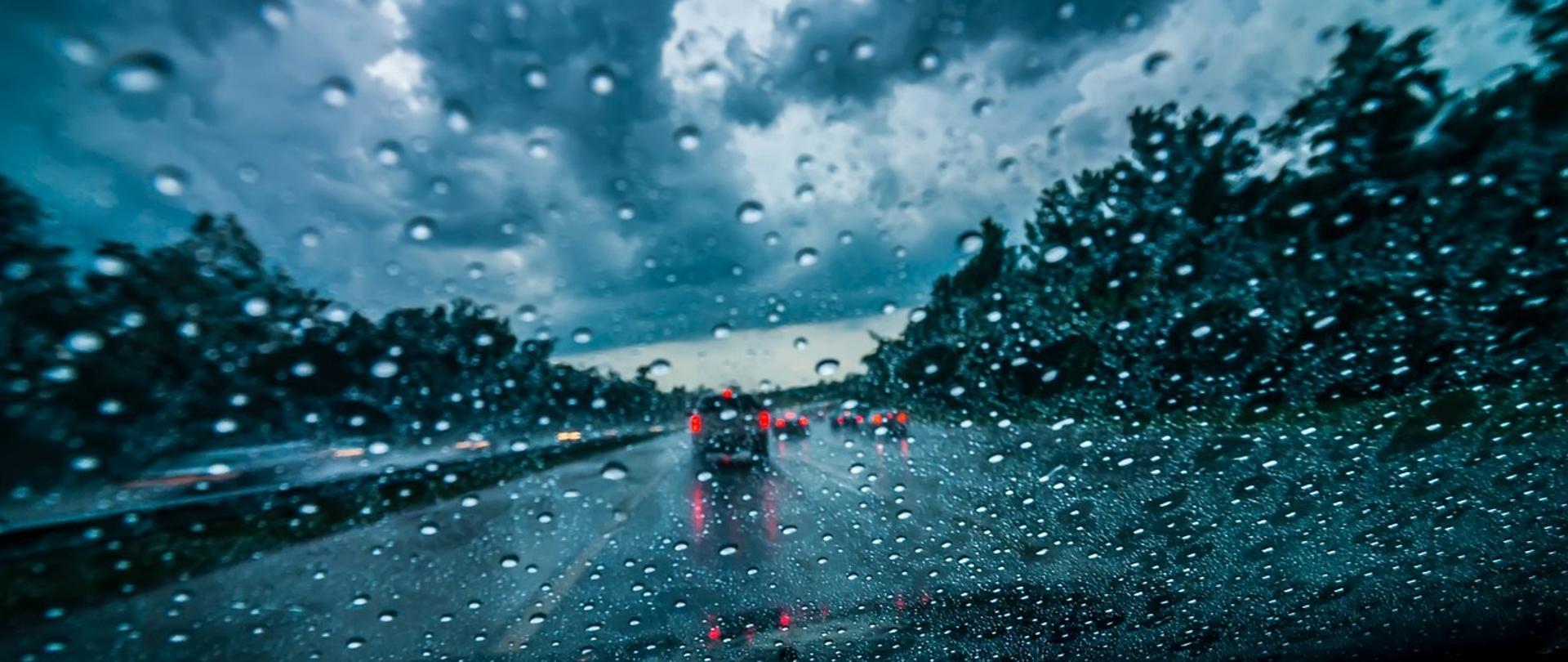 Zdjęcie przedstawia jezdnię, z perspektywy kierowcy samochodu, przez zachlapaną deszczem przednią szybę.