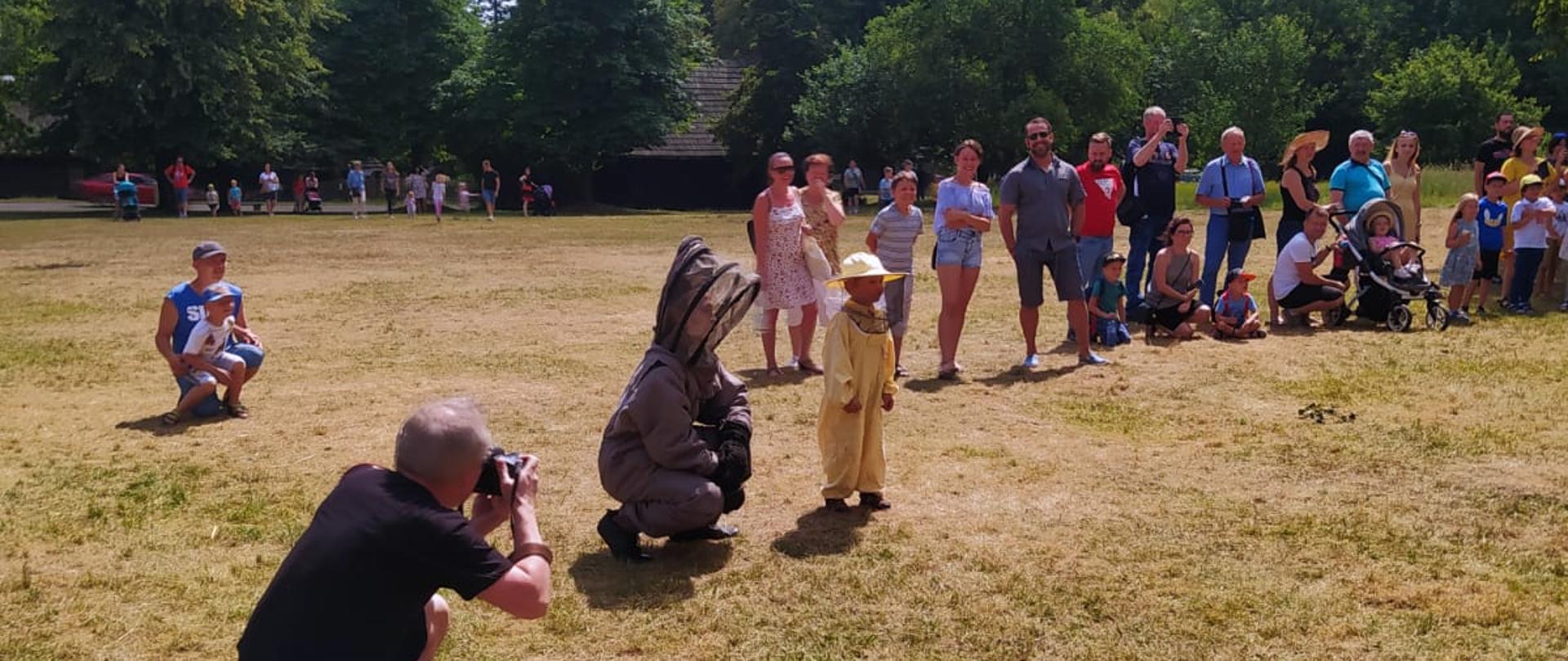 Zdjęcie przedstawia kokon pszczół wiszący na drzewie do którego jest rozstawiana drabina mechaniczna z Państwowej Straży Pożarnej. Przed drzewem stoją uczestnicy wydarzenia robiący zdjęcia. 