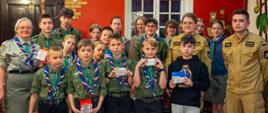 Zdjęcie zbiorowe, z lewej strony druhna harcmistrz w szarym mundurze harcerskim, po prawej stronie dwoje strażaków w mundurach służbowych, w kolorze piaskowym. Pomiędzy nimi około 20 harcerek i harcerzy w mundurkach zielonych. Wszyscy w ręku trzymają czujkę dymu lub czadu. W tle okno, drzwi i ściana w kolorze czerwonym. 