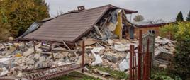 Widok zawalonego budynku po wybuchu gazu w Kaczorowie. Na pierwszym planie torba medyczna strażaków. Budynek praktycznie całkowicie zawalony, przykryty dachem, widoczna jedna niezawalona ściana. Uszkodzone ogrodzenie, w tle budynek z cegły. 