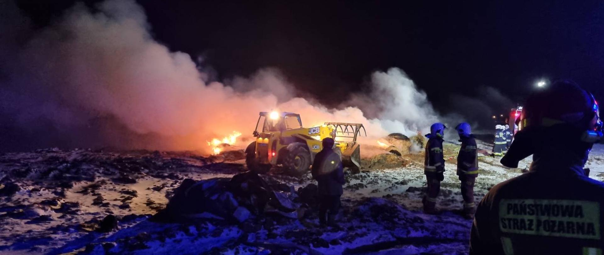 Zdjęcie przedstawia palący się stóg słomy w balotach. Na zdjęciu widać również ładowarkę, która rozgarnia palące się baloty w celu przelania wodą. Na zdjęciu widać również strażaków. Pora nocna. 
