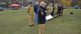 Warszaty szkoleniowe. Zdjęcie przedstawia fragment stadionu w Sarnowie. na pierwszym planie zastępca komendanta powiatowego PSP w Rawiczu, który przemawia przez mikrofon, trzymany w ręce. Za nim, w szeregu stoi kilkunastu mężczyzn - instruktorów, którzy będą prowadzić zajęcia. Część z nich to strażacy w ubraniach specjalnych koloru piaskowego i mundurach koszarowych. W tle drzewa, samochody i czerwony namiot. 