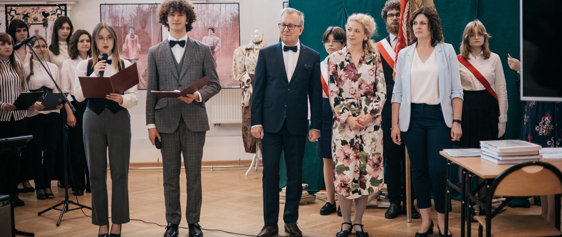 Zdjęcie przedstawia uroczystość rozdania dyplomów. W centralnej części zdjęcia znajduje się dyrekcja i uczniowie szkoły, w tle zdjęcia widoczne są prace dyplomantów.