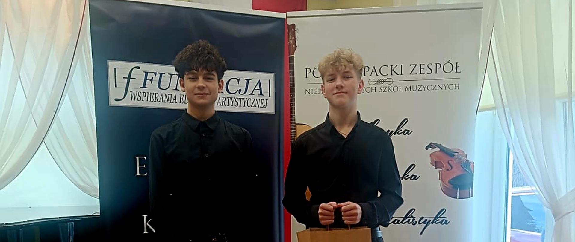 Dwóch uczniów ubranych na czarno, stoi na tle banerów, w tle okna, światło dnia świeci przez okno.