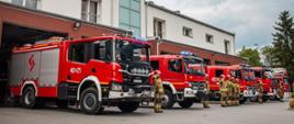 Na zdjęciu widać na kostce brukowej stoi pięć ciężarowych pojazdów pożarniczych w kolorze czerwonym z włączonymi światłami drogowymi. W jednym pojeździe widać włączone światło błyskowe niebieskie. Przy każdym samochodzie po prawej stronie stoi strażak ubrany w piaskowy mundur specjalny na głowach maja założone czerwone hełmy i salutują. Miedzy drugim a trzecim pojazdem stoją w szeregu czterech strażaków ubranych w piaskowe mundury i czerwone hełmy pierwszy z nich dowódca salutuje. Za strażakami widać budynek Komendy Powiatowej PSP w Bochni.