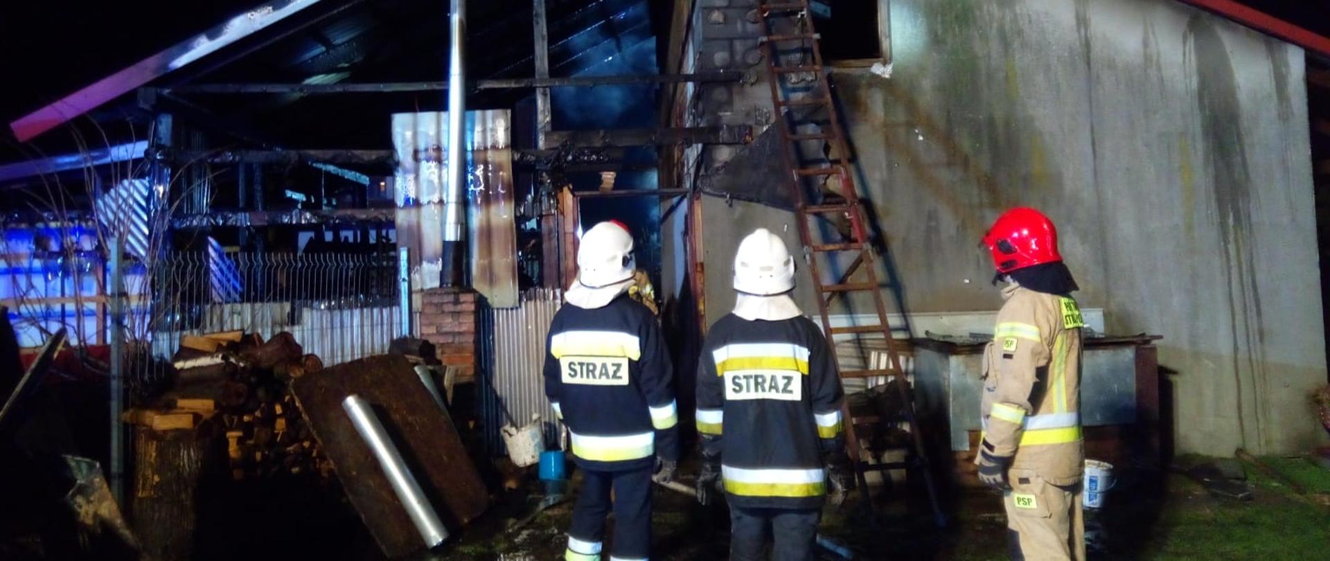 Zdjęcie przedstawia budynek gospodarczy w którym wybuchł pożar. Przed budynkiem stoi 3 ratowników a do ściany budynki dostawiona jest drabina. 