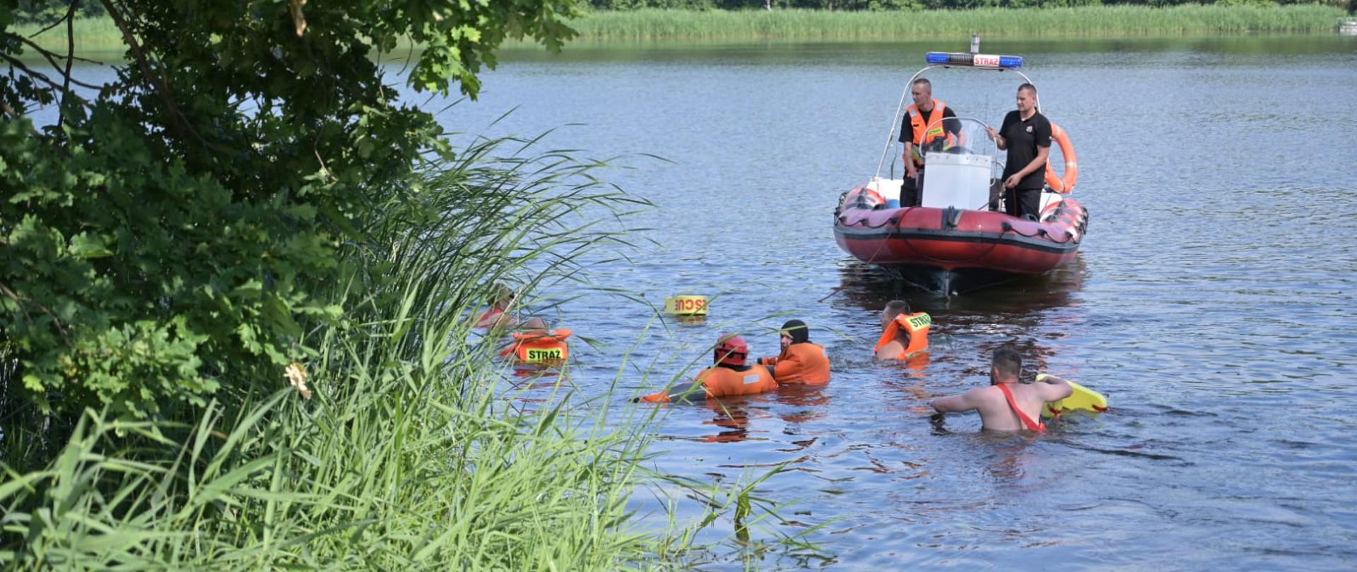 Zdjęcie przedstawia jezioro, po którym pływa czerwona strażacka łódź. Przed nią w wodzie znajdują się mężczyźni w kamizelkach asekuracyjnych i kombinezonach wypornościowych. W tle przybrzeżna roślinność.