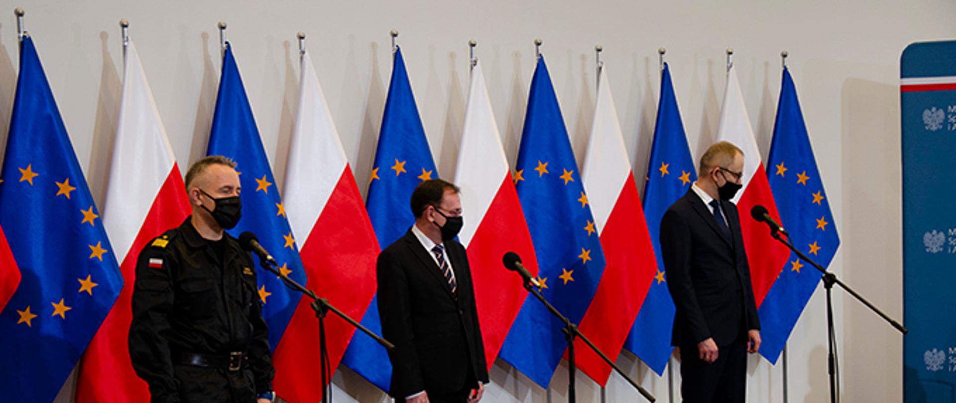 Służby ratownicze oraz przedstawiciele MSWiA biorące udział w konferencji stoją przed mikrofonami, w tle flagi unii europejskiej i Polski