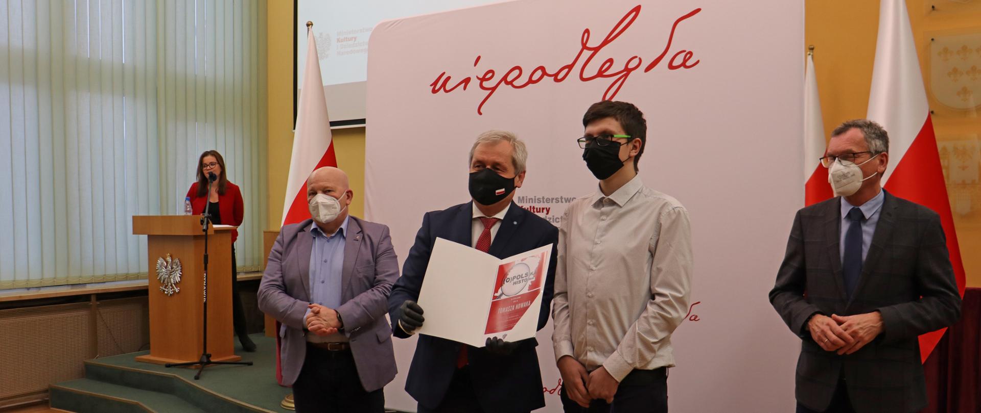 Na zdjęciu 5 osób. 4 z nich znajduje się w bliskiej odległości od obiektywu. Na środku kadru stoi wojewoda opolski, który pozuje do zdjęcia ze stojącym obok niego młodym człowiekiem - laureatem konkursu historycznego. Wojewoda trzyma w dłoni tęczkę z dyplomem konkursowym. Obok stoi wicedyrektor Instytutu Śląskiego oraz kurator oświaty. W tle widać ściankę z napisem Niepodległa oraz drewnianą mównicę z orzełkiem, za którą stoi prowadząca uroczystość.