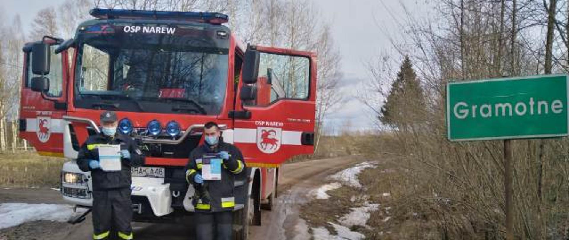 Widoczny pojazd gaśniczy uczestniczący w kolportażu ulotek na terenie gminy Narew, droga żwirowa, ratownicy z ulotkami na tle samochodu gaśniczego i tablicy z miejscowością Gramotne