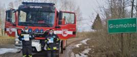 Widoczny pojazd gaśniczy uczestniczący w kolportażu ulotek na terenie gminy Narew, droga żwirowa, ratownicy z ulotkami na tle samochodu gaśniczego i tablicy z miejscowością Gramotne