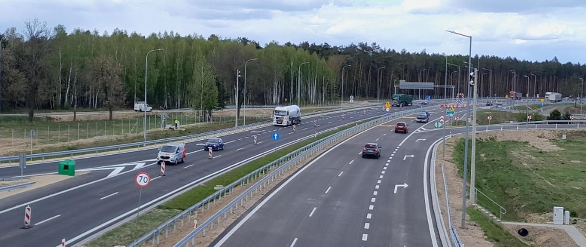 Widok z góry na nową, dwujezdniową drogę. Po jezdniach poruszają się samochody osobowe i ciężarowe. Widoczne są pasy rozdzielające, oznakowanie poziome i pionowe, ograniczenie prędkości do 70 km/h oraz elementy infrastruktury drogowej. W tle las i tereny zielone.