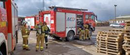 Na pierwszym planie widać strażaków zakładających sprzęt ochrony układu oddechowego. W tle stoją samochody pożarnicze na terenie przedsiębiorstwa. 