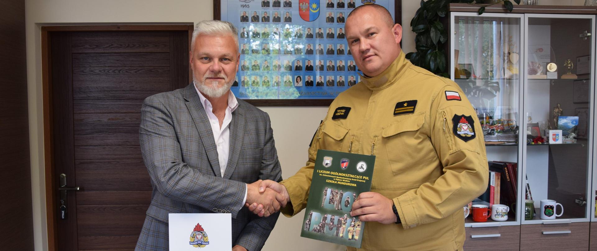 Komendant Powiatowy PSP w Mińsku Mazowieckim oraz Dyrektor Szkoły I LO PUL w Dębem Wielkim po podpisaniu porozumienia o współpracy. Stoją na tle ściany, podają sobie ręce. Od lewej Dyrektor Szkoły i Komendant