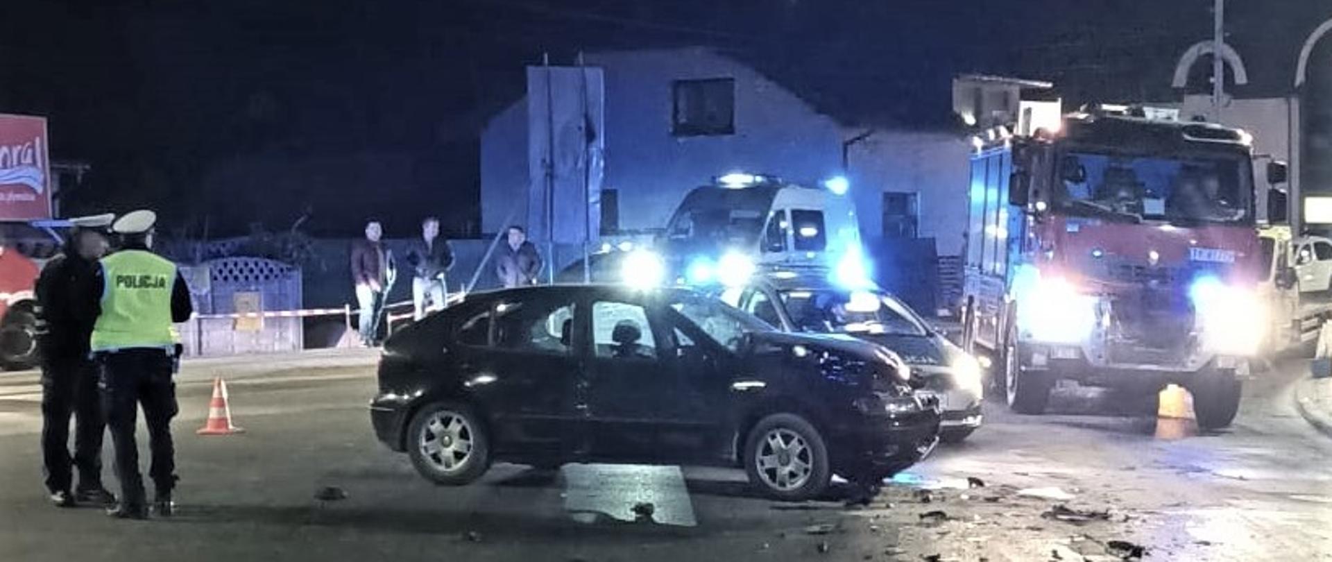 Na zdjęciu przedstawiono miejsce wypadku: na pierwszym planie uszkodzony Seat, przy nim stoją dwaj funkcjonariusze Policji. Na dalszym planie samochody służb ratowniczych. 