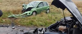 Na zdjęciu widać dwa samochody osobowe po zderzeniu czołowym. Oby dwa samochody maja rozwalony przód pojazdu. Jeden z nich znajduje się na drodze powiatowej, a drugi na poboczu.
