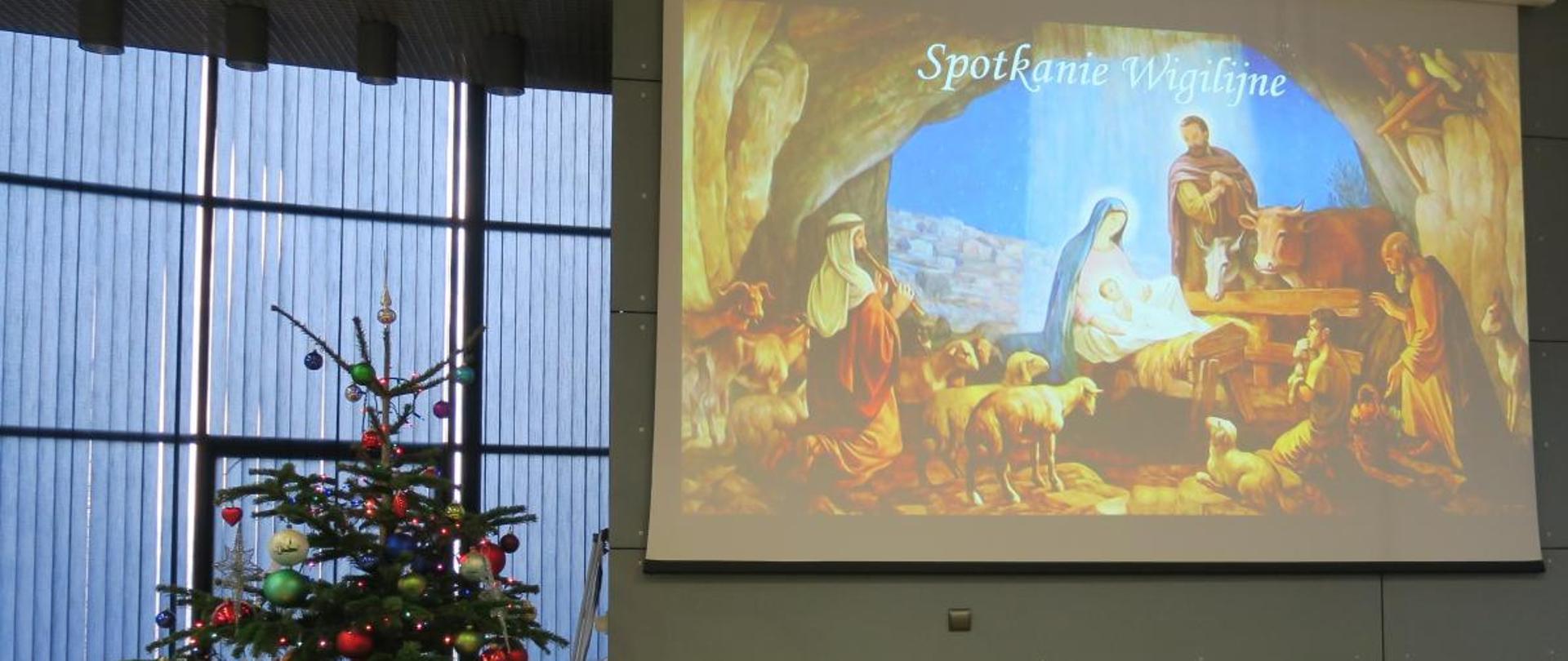 Po lewej stronie choinka przyozdobiona lampkami i bombkami, po prawej na ścianie wyświetlona szopka Bożonarodzeniowa.