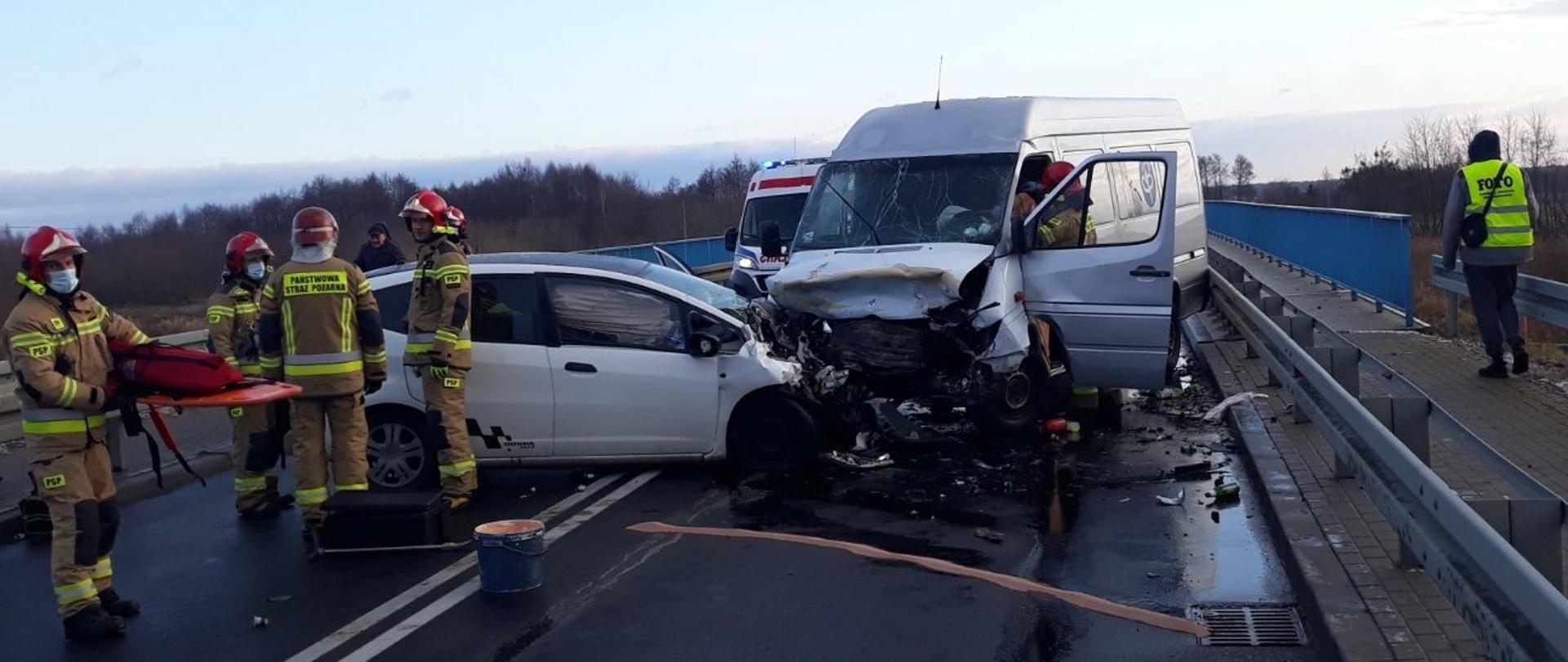 Na jezdni dwa samochody, w tym jeden osobowy, drugi dostawczy, po zderzeniu. Obok samochodów leżą pozostałości uszkodzonych elementów. Przed pojazdami stoją strażacy. Jeden z nich trzyma deskę ortopedyczną przygotowana do użycia. Przez otwarte drzwi samochodu dostawczego strażak zaopatruje poszkodowanego kierowcę. Za pojazdami stoi karetka pogotowia. 