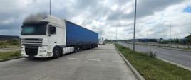Na pierwszym planie (od lewej): ciągnik siodłowy z podpiętą naczepą kurtynową typu firanka. Za kontrolowanym zespołem pojazdów stoi oznakowany furgon mazowieckiej Inspekcji Transportu Drogowego. Pojazdy znajdują się w zatoce kontrolnej przy dwupasowej drodze krajowej numer 9 w Radomiu. W tle: w oddali widać dużą halę produkcyjną pobliskiego zakładu i inne zabudowania.