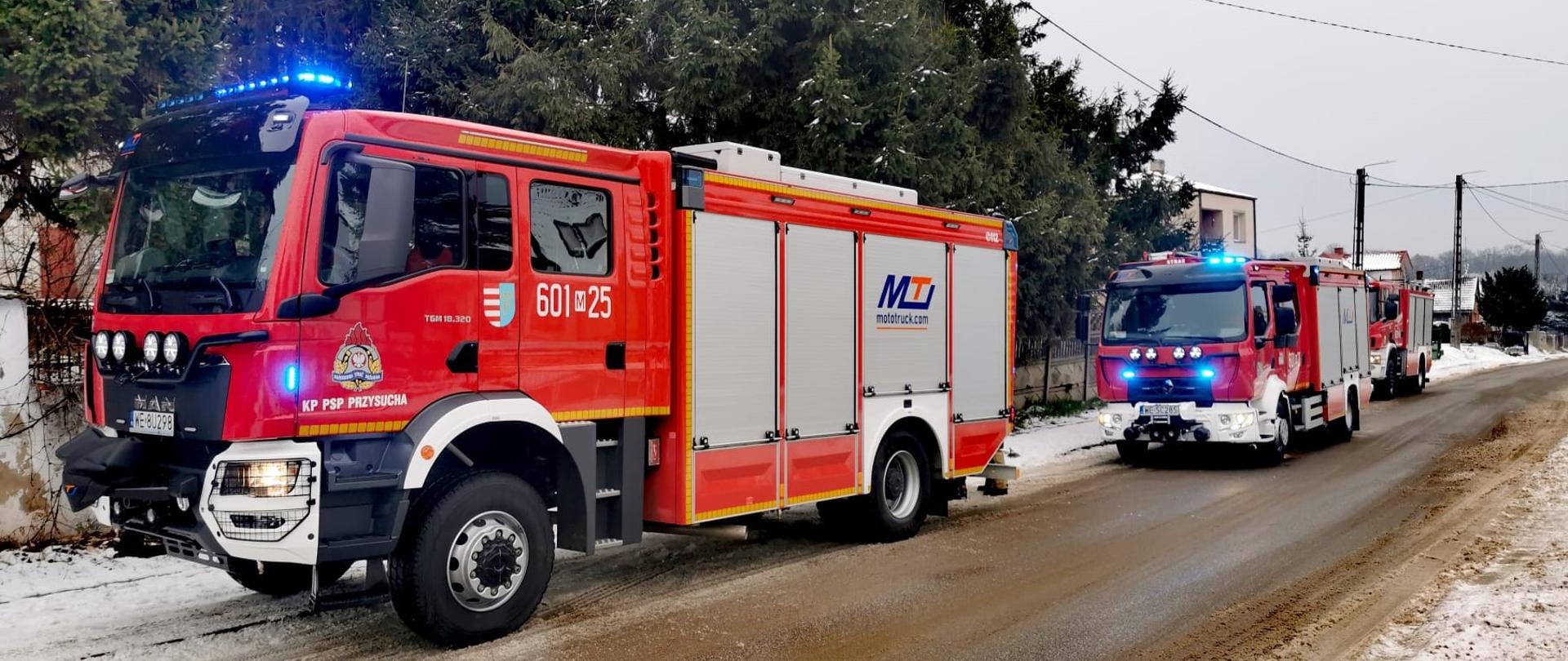 Zdjęcie przedstawia kolumnę trzech czerwonych wozów strażackich jadących zaśnieżoną, częściowo pokrytą błotem pośniegowym drogą. Jest to zimowy dzień, a po bokach drogi widać leżący śnieg. Na pierwszym planie, po lewej stronie, znajduje się duży wóz strażacki z włączonymi niebieskimi światłami awaryjnymi (kogutami) na dachu. Na drzwiach kabiny widoczny jest numer "601 25" oraz napis "KP PSP PRZYSUCHA". Jest to nowoczesny pojazd gaśniczy.
Za pierwszym wozem, w oddali, widać kolejny, nieco mniejszy wóz strażacki, również z włączonymi niebieskimi światłami. Na jego boku widoczne jest logo "MTI". W tle po lewej stronie znajdują się drzewa iglaste oraz ogrodzenie. Po prawej stronie drogi widoczne są słupy energetyczne i fragmenty budynków w oddali. Cała scena sugeruje, że wozy strażackie są w drodze na interwencję lub wracają z niej, poruszając się w zimowych warunkach. 