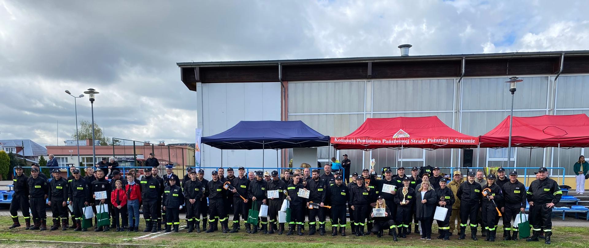 pora dzienna, pochmurno. Na boisku ustawienie są uczestnicy i goście zawodów OSP. Z tyłu widać rozstawione namioty.