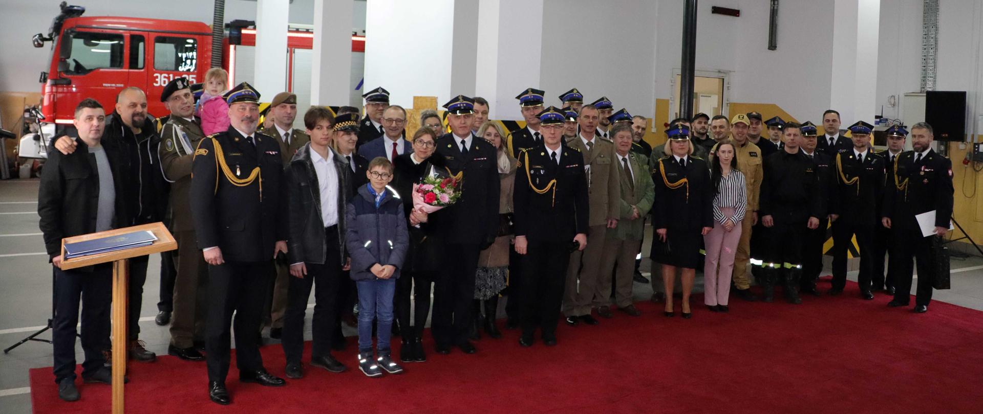 Zdjęcie przedstawia grupowe zdjęcie uczestników uroczystej zbiórki z okazji odejścia na zaopatrzenie emerytalne st. bryg. Grzegorza Lisika. Zdjęcie zostało wykonane w garażu na tle pojazdu pożarniczego.