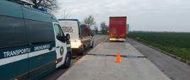 Pojazdy Inspekcji Transportu Drogowego podczas kontroli drogowej. Na poboczu ustawione są dwa oznakowane pojazdy ITD, a na jezdni znajduje się waga do pomiaru masy pojazdów oraz pachołek ostrzegawczy. W tle widoczna jest ciężarówka oraz rozległe pola uprawne pod pochmurnym niebem.