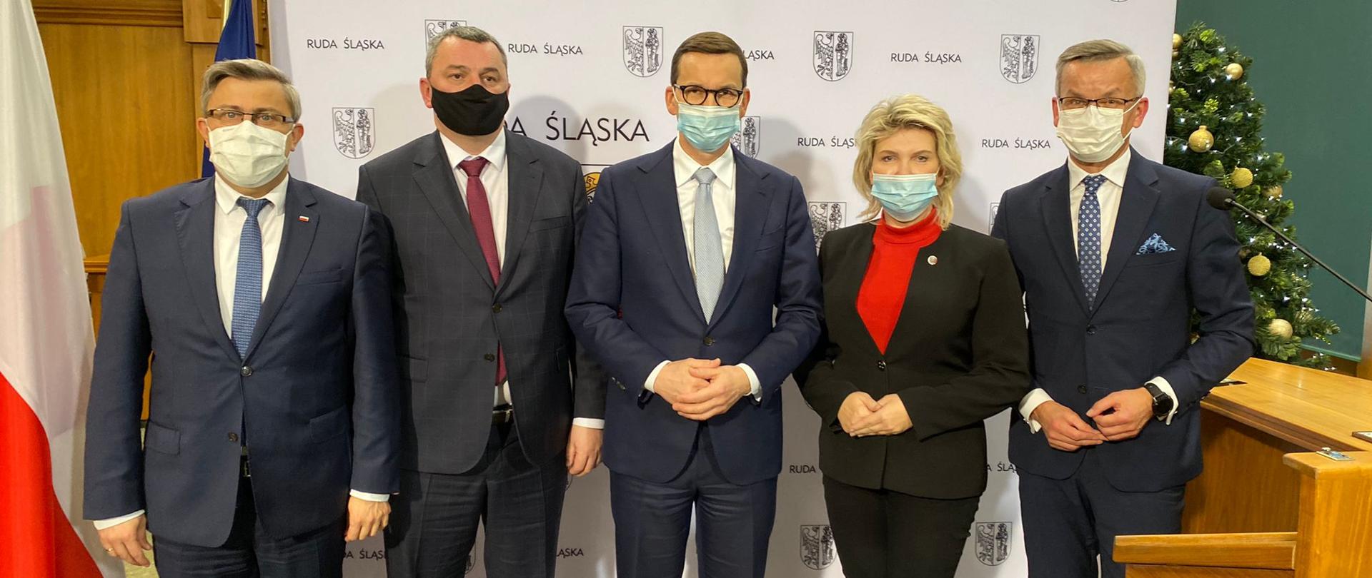 Na tle herbu oraz napisów Ruda Śląska od lewej stoją: Wojewoda Śląski Jarosław Wieczorek, Poseł na Sejm RP Marek Wesoły, Premier Mateusz Morawiecki, Senator RP Dorota Tobiszowska, Zastępca Prezydenta Miasta Ruda Śląska Krzysztof Mejer