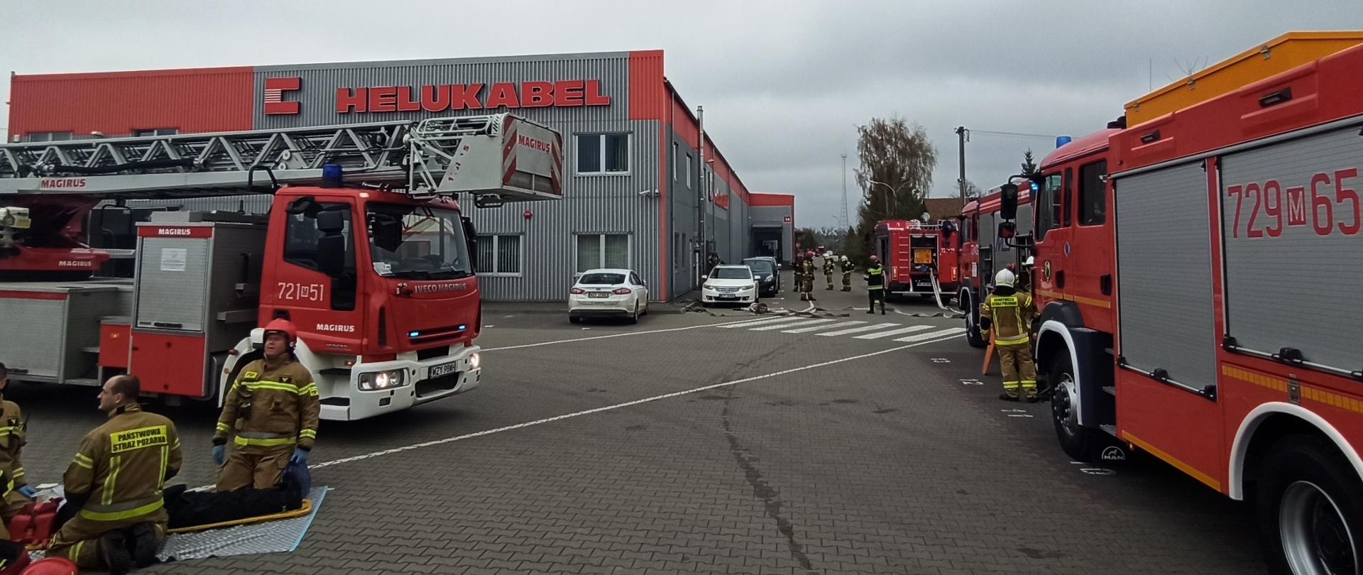 Samochody pożarnicze stoją przed budynkiem zakładu Helukabel. Po lewej stronie przed samochodem pożarniczym na macie, przy osobie poszkodowanej klęczą strażacy.